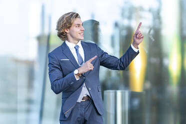 Cheerful young businessman gesturing against glass office building - GGGF00884