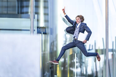 Cheerful male entrepreneur with laptop gesturing while jumping against glass office building - GGGF00887