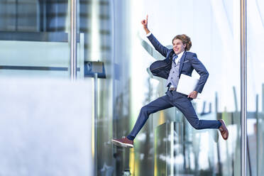 Fröhlicher männlicher Unternehmer mit Laptop, der gestikulierend gegen ein gläsernes Bürogebäude springt - GGGF00887