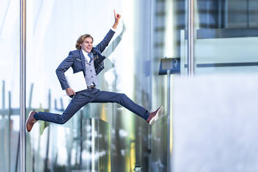 Aufgeregter männlicher Berufstätiger mit Laptop, der beim Sprung gegen ein Bürogebäude zeigt - GGGF00888