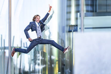 Excited male professional with laptop pointing while jumping against office building - GGGF00888