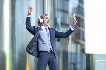 Fröhlicher junger Geschäftsmann hört Musik, während er ein Selfie gegen Glas macht - GGGF00889