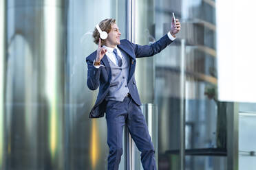 Glückliche männliche professionelle Geste bei der Aufnahme von Selfie durch Smartphone gegen Bürogebäude - GGGF00890