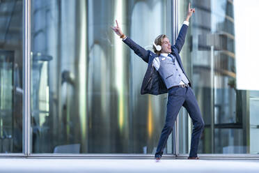 Cheerful young businessman with arms raised dancing while listening music against glass - GGGF00892