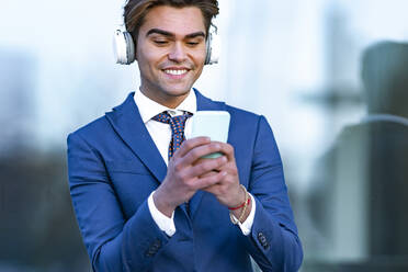 Happy male professional using smart phone while listening music against office building - GGGF00894