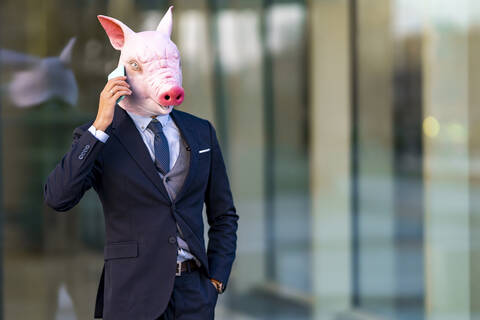 Young businessman in pig mask with hands in pockets talking on mobile phone against glass stock photo