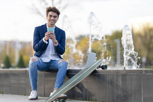 Lächelnder junger Geschäftsmann mit Skateboard und Smartphone, der auf einer Stützmauer sitzt - GGGF00918