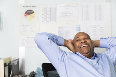 Cheerful employee with hands behind head laughing while sitting in print shop - ISPF00026