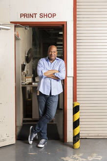Smiling supervisor with arms crossed leaning on doorway at print shop - ISPF00027