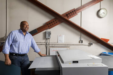 Happy supervisor looking away while standing by printer in print shop - ISPF00030