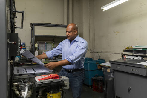 Male employee examining paper by print machine in shop - ISPF00036