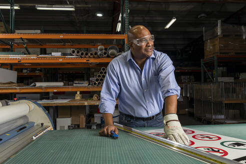 Smiling employee wearing protective eyewear looking away while working in print shop - ISPF00038