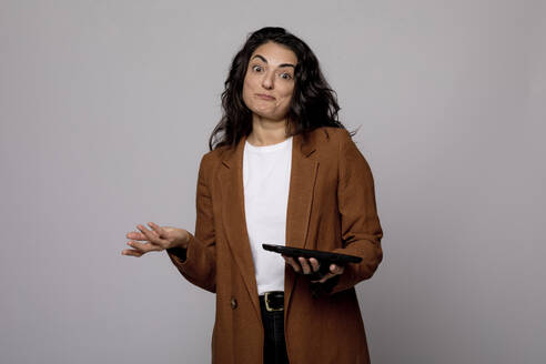 Confused woman with digital tablet gesturing against gray background - FLLF00562