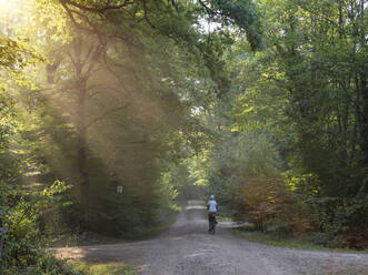 Senior female biker riding E-bike on dirt road in green forest - LAF02632