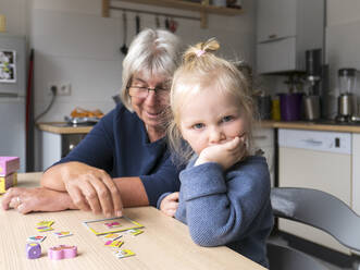 Upset blond girl sitting by grandmother playing puzzle at dining table in kitchen - LAF02633