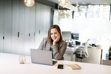 Smiling businesswoman with hand on chin using laptop at desk in office - JCZF00444