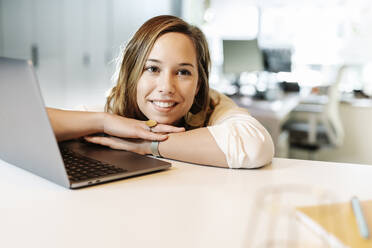 Close-up of smiling businesswoman contemplating while leaning by laptop on desk - JCZF00446