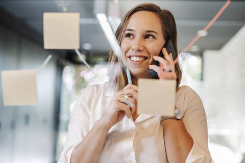 Close-up of smiling businesswoman talking over smart phone in office seen through glass wall stock photo