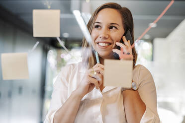 Nahaufnahme einer lächelnden Geschäftsfrau, die über ein Smartphone im Büro spricht, gesehen durch eine Glaswand - JCZF00457