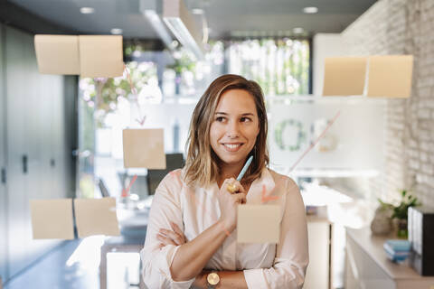 Smiling businesswoman looking at adhesive notes stuck on glass wall in office stock photo