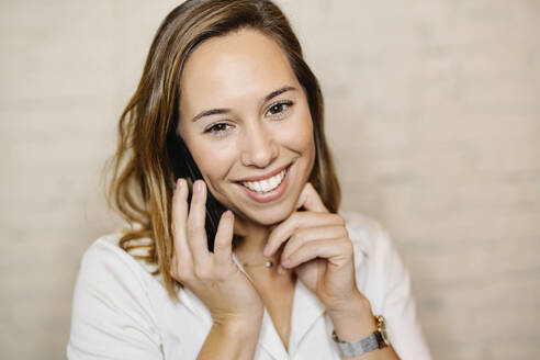 Nahaufnahme einer lächelnden weiblichen Fachkraft, die über ein Smartphone an der Wand spricht - JCZF00466
