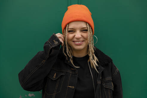 Glückliche Hipster-Frau mit geflochtenem Haar vor grüner Wand - ACPF01024