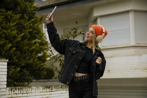 Junge Frau mit Strickmütze nimmt Selfie durch Smartphone gegen Gebäude, lizenzfreies Stockfoto