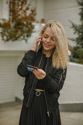 Blond young woman looking at credit card while talking on smart phone in front yard - ACPF01050