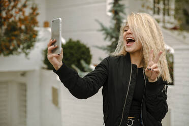 Blonde junge Frau macht Frieden Geste während der Einnahme selfie durch Handy - ACPF01052
