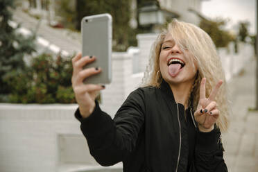 Glückliche junge Frau, die ein Selfie macht, während sie friedlich gestikuliert und die Zunge durch ihr Smartphone herausstreckt - ACPF01053
