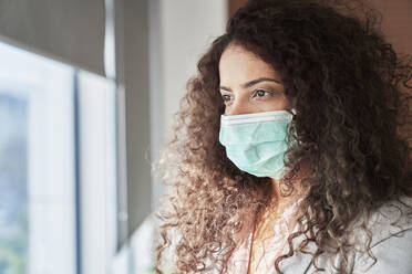 Female professional wearing protective face mask looking through window from office - ABIF01300