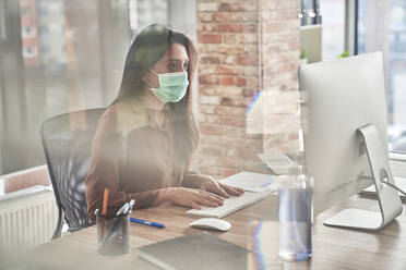 Mid adult woman working on computer on desk seen through glass during pandemic - ABIF01303