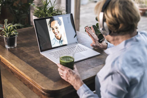 Grandmother waving to granddaughter on video call through laptop while having juice at home stock photo