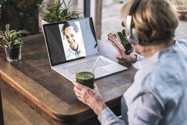 Grandmother waving to granddaughter on video call through laptop while having juice at home - UUF22655