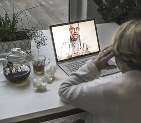 Ältere Frau führt ein Videogespräch mit einem männlichen Arzt über einen Laptop im Wohnzimmer - UUF22661
