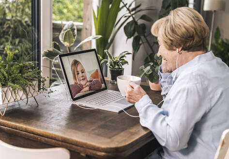 Lächelnde Großmutter bei einem Videogespräch mit ihrem Enkelkind über den Laptop, während sie zu Hause einen schwarzen Kaffee trinkt - UUF22675