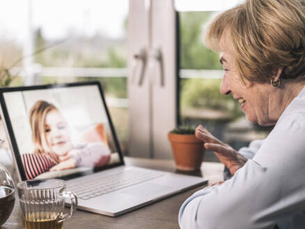 Lächelnde Großmutter, die ihrem Enkelkind bei einem Videogespräch über einen Laptop im Wohnzimmer zuwinkt - UUF22677