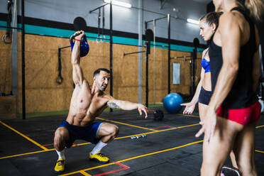 Männlicher Fitnesstrainer hebt eine Kettlebell und übt Kniebeugen, während er Frauen im Fitnessstudio hilft - MIMFF00422