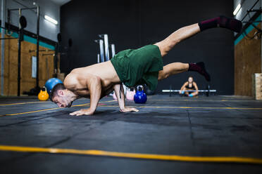 Hemdloser junger Mann balanciert auf dem Boden, während die Frau im Hintergrund im Fitnessstudio ruht - MIMFF00425