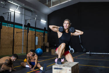 Eine Sportlerin hebt Hanteln, während sie auf einer Box trainiert, und ein Mann ruht sich im Fitnessstudio aus - MIMFF00427