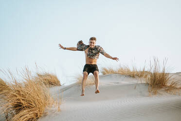 Junger Mann mit aufgeknöpftem Hemd, der auf Sand in Almeria, Tabernas-Wüste, Spanien, springt - MIMFF00442