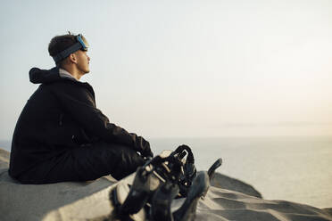 Junger Mann mit Schutzbrille sitzt auf einem Sandbrett in der Wüste von Almeria, Tabernas, Spanien - MIMFF00449