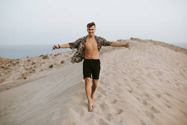 Fröhlicher junger Mann, der das Shaka-Zeichen macht, während er auf Sanddünen in Almeria, Tabernas-Wüste, Spanien, spazieren geht - MIMFF00451