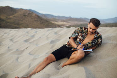 Gut aussehender Mann, der in ein Buch schreibt, während er auf dem Sand in Almeria, Tabernas-Wüste, Spanien, sitzt - MIMFF00453