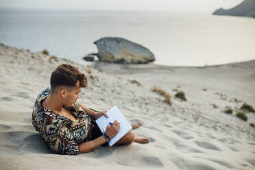 Junger Mann sitzt auf einer Sanddüne und zeichnet in einem Buch in Almeria, Tabernas-Wüste, Spanien - MIMFF00454