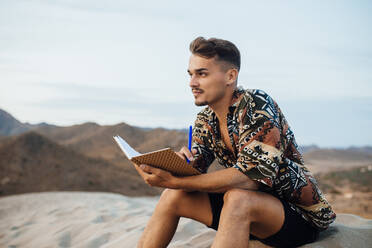 Young man sitting on sand dunes looking away while holding book and pen against sky at Almeria, Tabernas desert, Spain - MIMFF00456