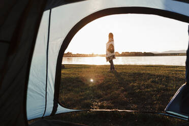 Woman wrapped in shawl standing on grassy land against lake seen through tent - EBBF02201