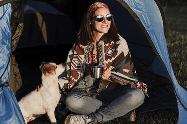 Glückliche Frau mit Sonnenbrille, die eine Tasse hält, während sie mit ihrem Hund im Zelt sitzt - EBBF02202