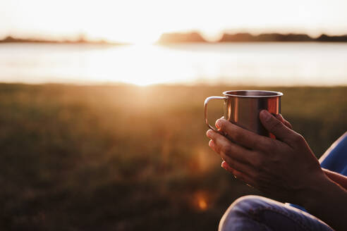 Hände einer mittleren erwachsenen Frau, die ein Getränk gegen einen See bei Sonnenuntergang hält - EBBF02205