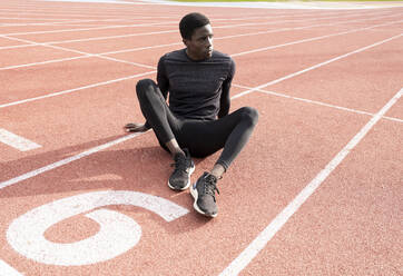 Sportsman contemplating while sitting on running track during sunny day - JCCMF00868